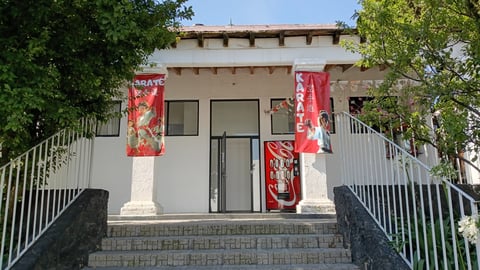 Beige building with red decorative banners, white railings, stone steps, and surrounding green trees and foliage