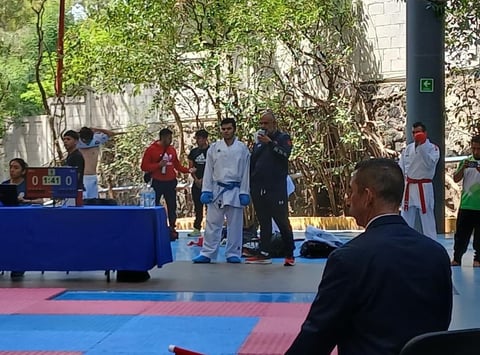 Martial arts competition with athletes in gi standing on a blue and pink mat, spectators watching from the sidelines in an outdoor courtyard setting.