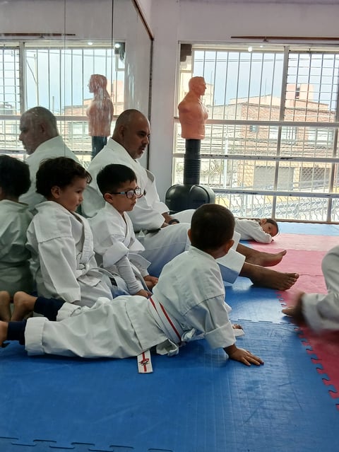 Children in white martial arts uniforms sitting on blue and pink exercise mats in a bright gym with windows overlooking a desert landscape