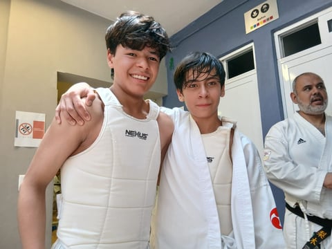 Two young people in white athletic uniforms posing together with an adult instructor visible in the background of a martial arts studio