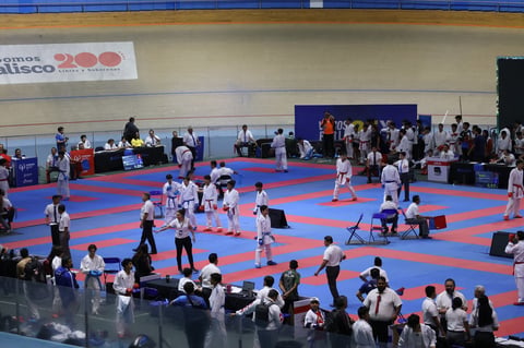 Karate or martial arts competition in a large indoor gymnasium with blue and red padded mats, athletes competing, and spectators watching from the sidelines.
