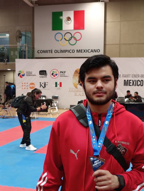Young athlete in red jacket holding medal at karate competition with Mexican Olympic Committee banner and blue mat in background