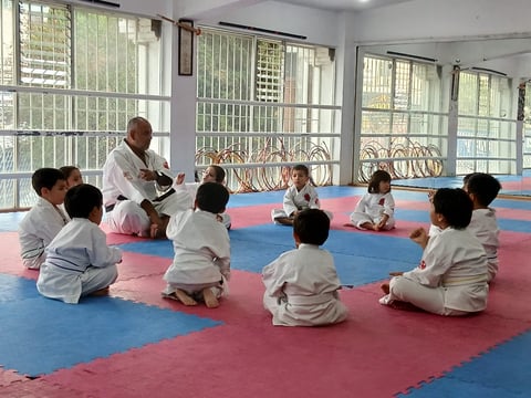 Instructor teaching group of young children in martial arts uniforms sitting on red and blue padded mat in training room