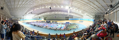 Indoor cycling arena with blue track, spectators in stands, and bright overhead lighting in large warehouse venue