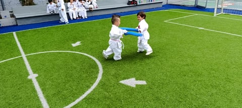 Two young children in white karate uniforms practice on an indoor green soccer field with white line markings