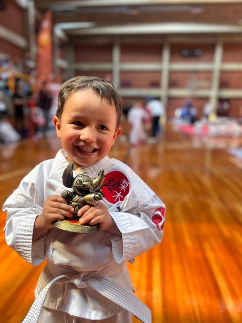 Young child in white karate gi holding trophy while smiling at camera in gymnasium
