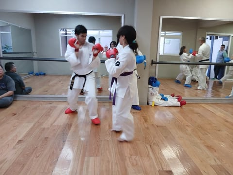 Two karate students in white uniforms and red gloves sparring in a mirrored studio with other students in background
