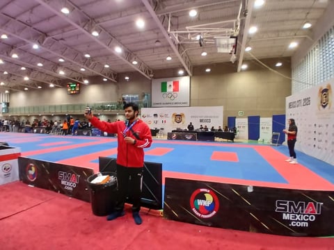 Athlete in red jacket standing on red mat at indoor martial arts competition venue with blue mats and Mexico flag signage