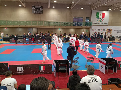 Karate competition at indoor gymnasium with athletes in white and pink gis on blue and red mat, spectators in foreground, Mexican flag visible on wall
