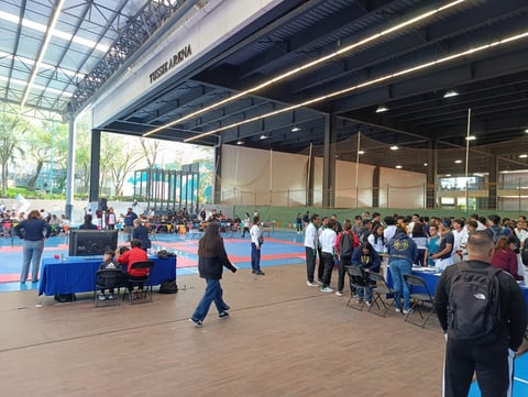 Large indoor sports facility with crowds of people gathered around blue mats, a green playground structure visible in background, and industrial ceiling with lighting.