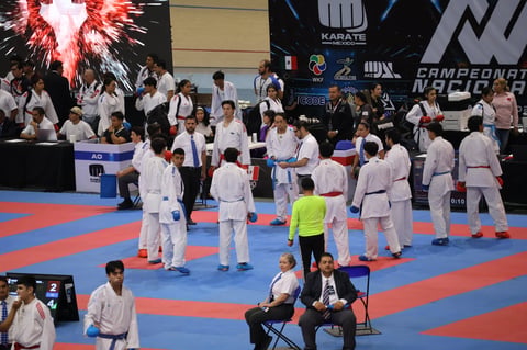 Karate competition venue with multiple athletes in white gis on a blue and red mat, spectators and officials visible in the background.