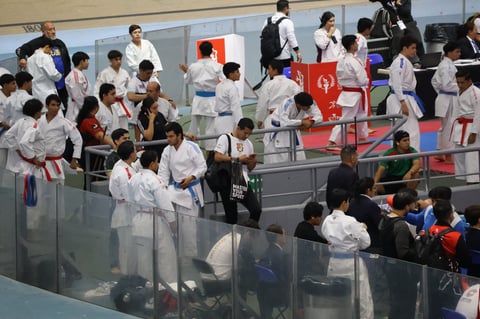 Martial arts competitors in white gis at an indoor tournament venue with spectators and judges observing from elevated areas.