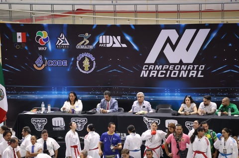 Panel of judges seated at table with large screen backdrop and competitors in white uniforms standing in front at what appears to be a martial arts championship event