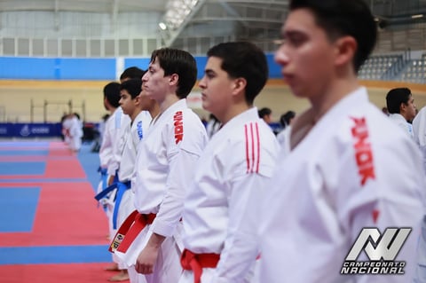 Young martial artists in white uniforms with red trim standing in a line at an indoor dojo or competition venue with blue and red mat