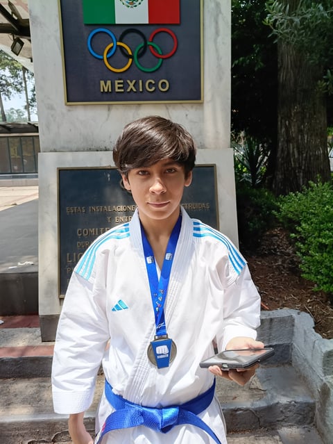 Young judoka in white gi and blue belt wearing medal in front of Mexico Olympic monument