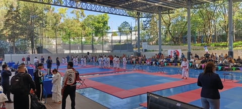 Outdoor martial arts tournament with participants competing on a red and blue mat under a metal pavilion with spectators watching.