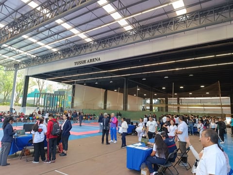 Large indoor arena with crowd of people standing on blue mat flooring, tables and spectators during what appears to be a sporting or community event