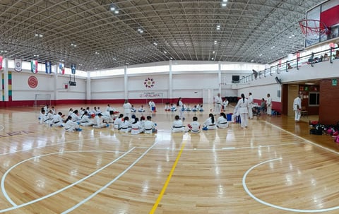 Group of students in white uniforms practicing martial arts or karate in a large indoor gymnasium with wooden flooring and court markings