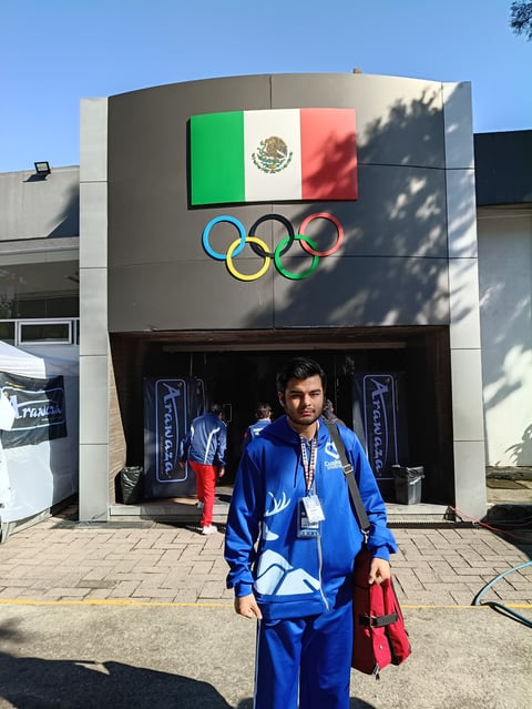 Person in blue athletic jacket standing in front of building with Mexican flag and Olympic rings symbol