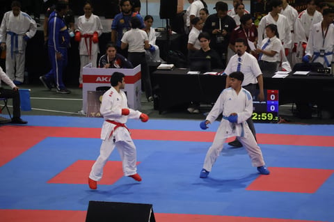 Two karate competitors in white gis facing off on a blue and red mat during a tournament competition with spectators and judges in background