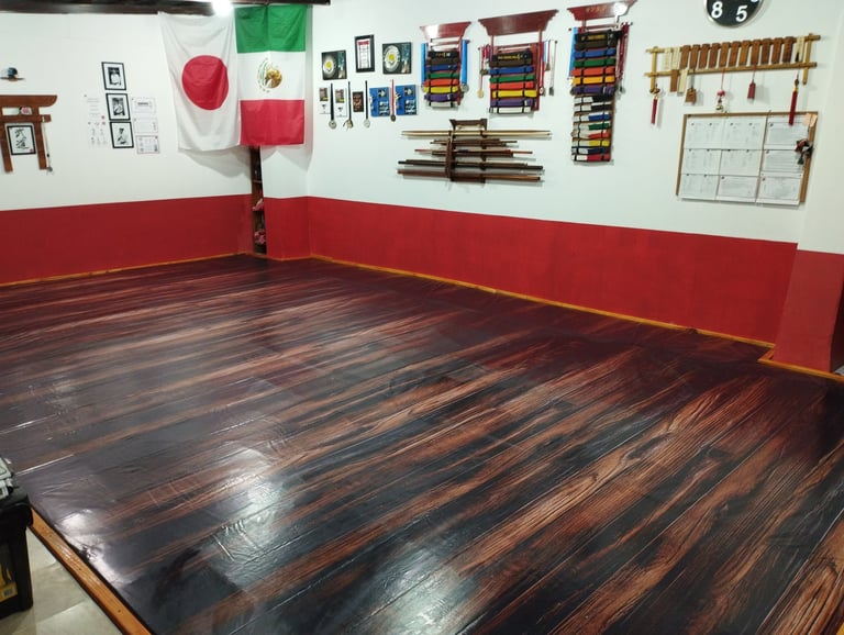 Martial arts training room with a large dark wooden mat, red and white walls, and organized equipment hanging on wall including targets, weapons racks, and training gear.